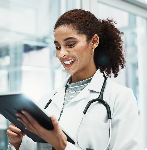 Smiling healthcare professional wearing a white coat and stethoscope, looking at a digital tablet in a bright clinical setting, representing modern patient care and technology for Bright Roads